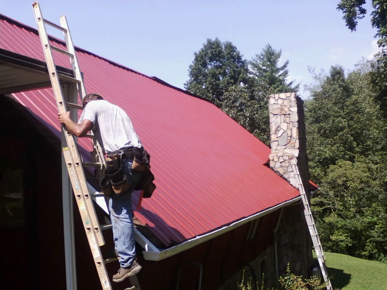 Roofer on a ladder beside a red metal roof and stone chimney — Soffit & Fascia Repair by Roofing Brothers in Clarkesville